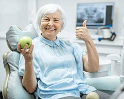 An older woman showing off her new dentures in Carrollton and giving a thumbs up while holding an apple