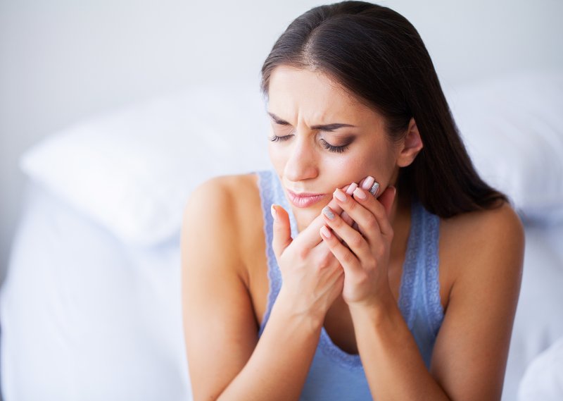 a woman seated on the side of her bed holding her cheek and cringing in pain